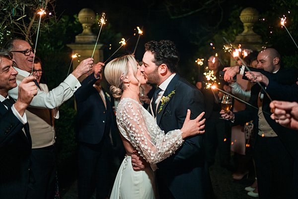 Bride and groom kiss surrounded by sparklers at night.
