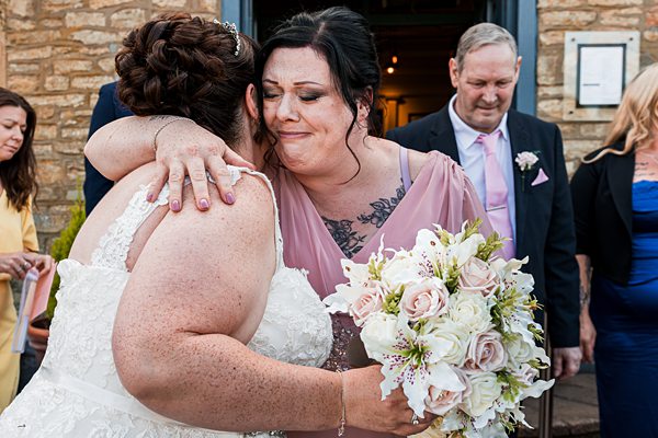 Two women hugging at a wedding ceremony, holding flowers.